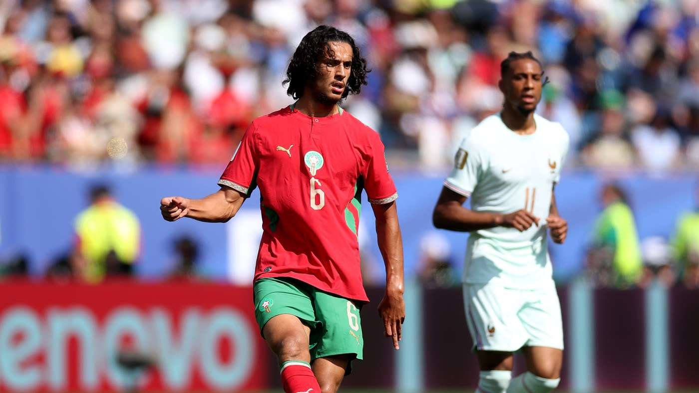 Soccer player in a red jersey with number 6 and green shorts runs on the field during a match, with a crowd in the stands behind him.