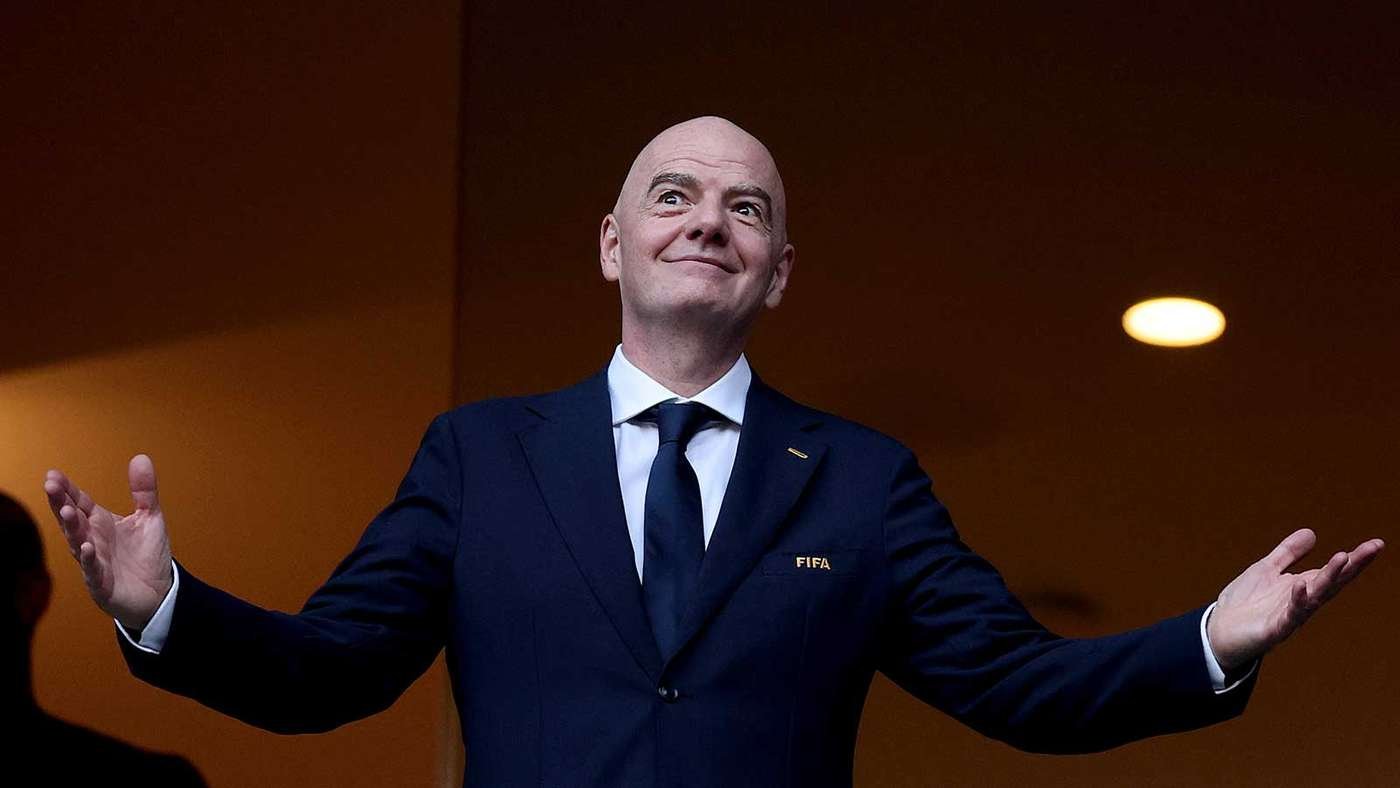 Smiling bald man in a navy suit with a FIFA badge and tie, arms outstretched in a welcoming gesture on stage.