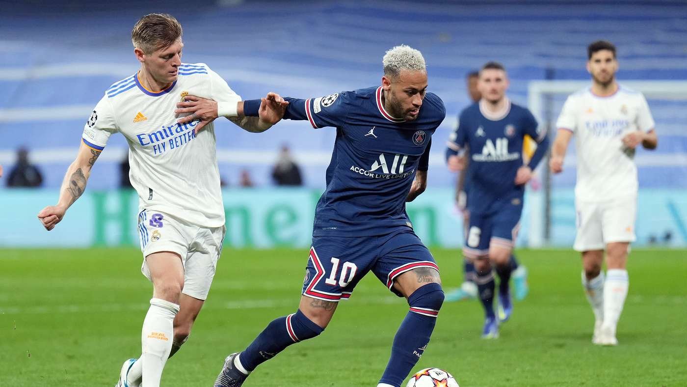 Soccer players contesting the ball during a match, Real Madrid in white versus PSG in navy kit on a green field.