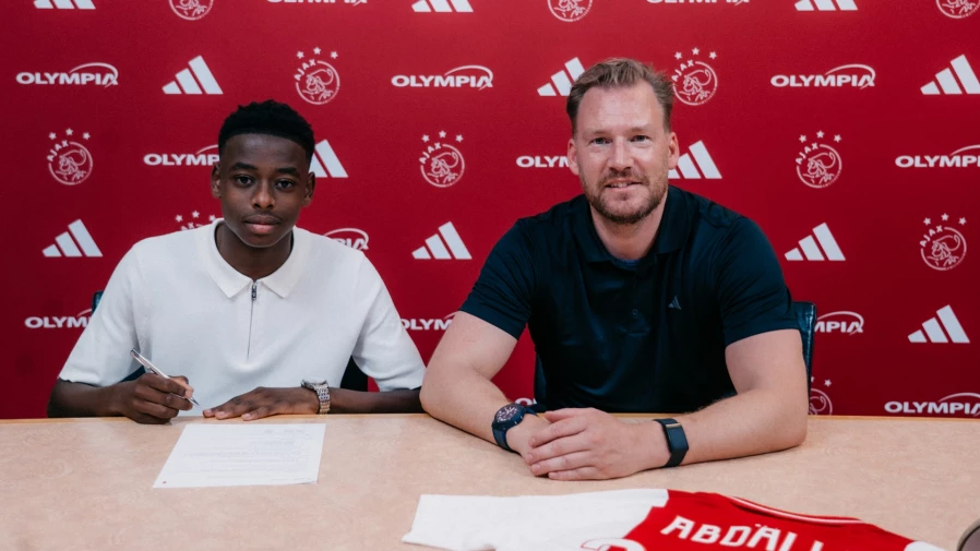 Young athlete signs a contract at a table with a coach beside him during a press event, red backdrop covered with Olympia logos.