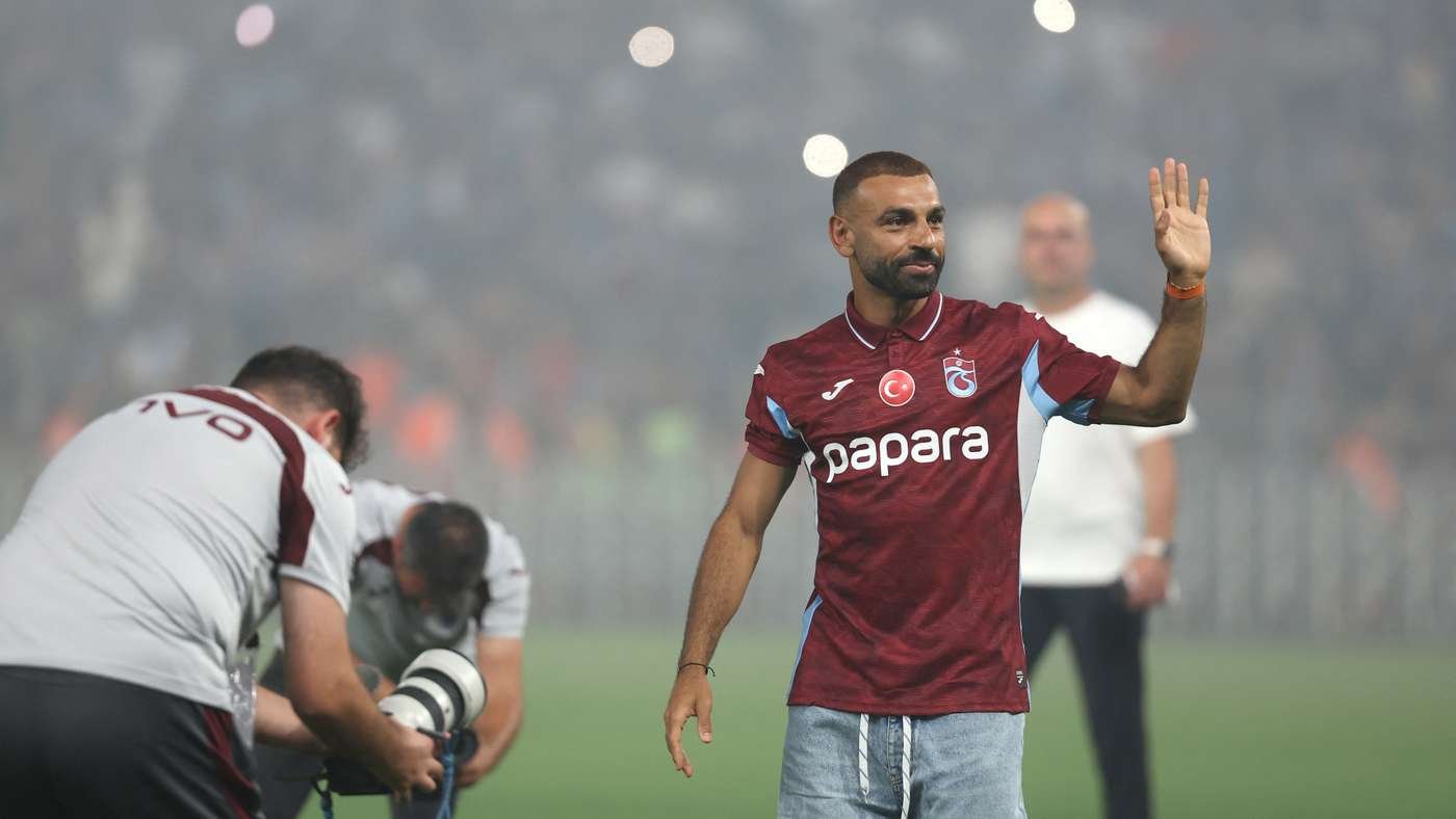 Soccer player in a maroon jersey waves to the crowd on a foggy field, with teammates and a photographer nearby.
