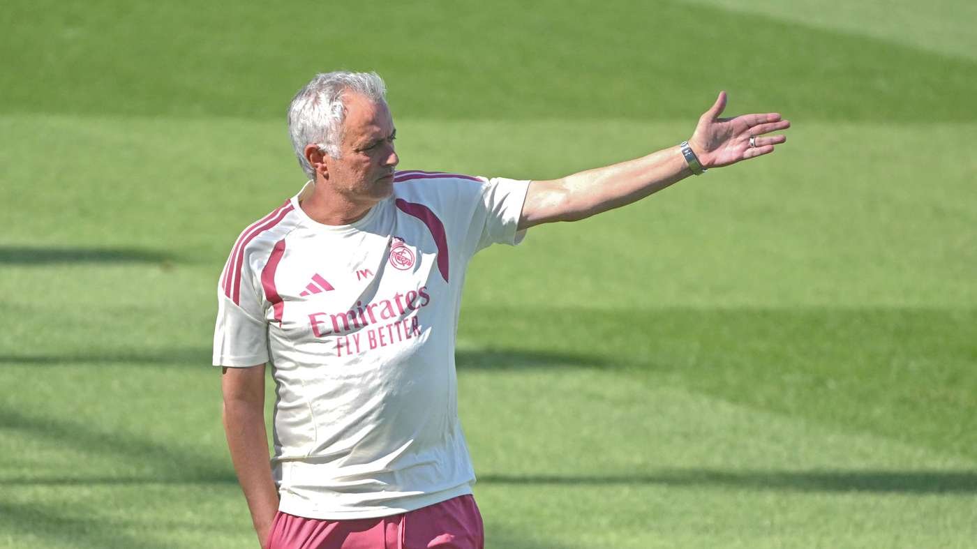 A man with gray hair in a Real Madrid training kit extends his right arm while standing on a green soccer field.