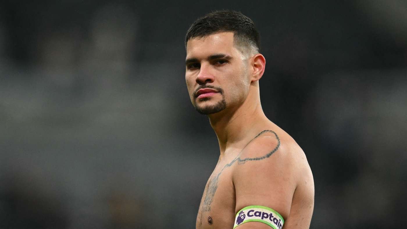 Male soccer player standing shirtless on the field, wearing a captain's armband, with a shoulder tattoo visible.