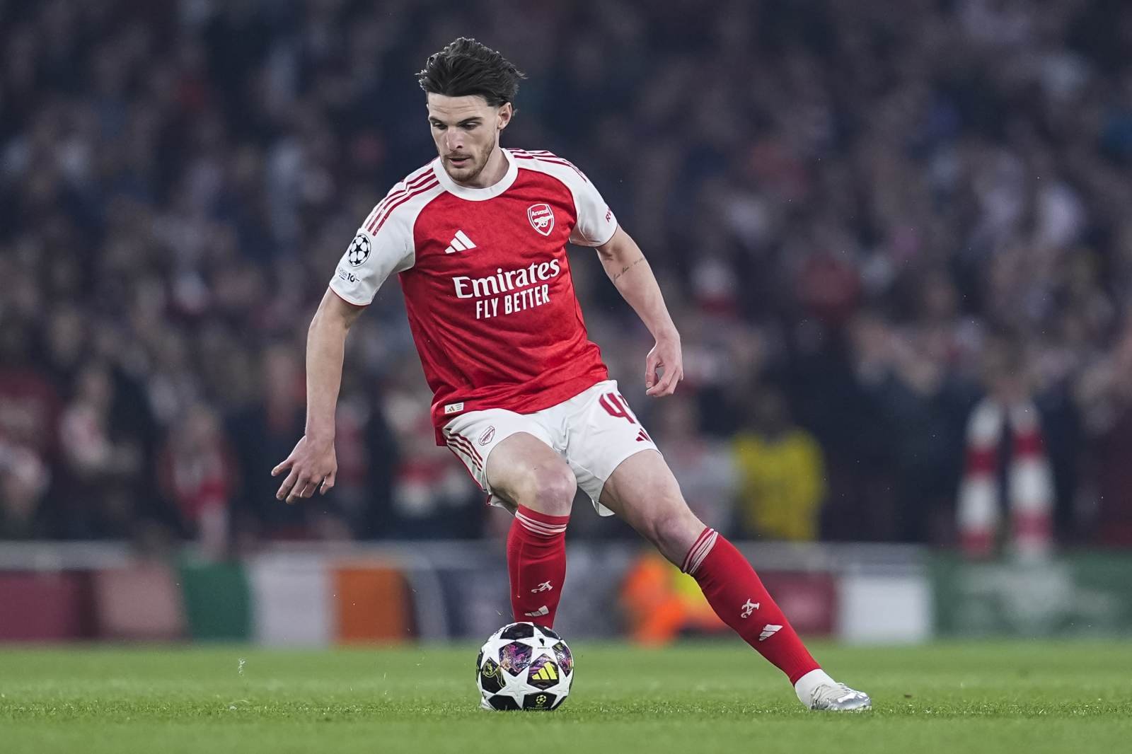 Arsenal player in red and white kit dribbling a soccer ball on a green pitch during a match with a crowd in the background