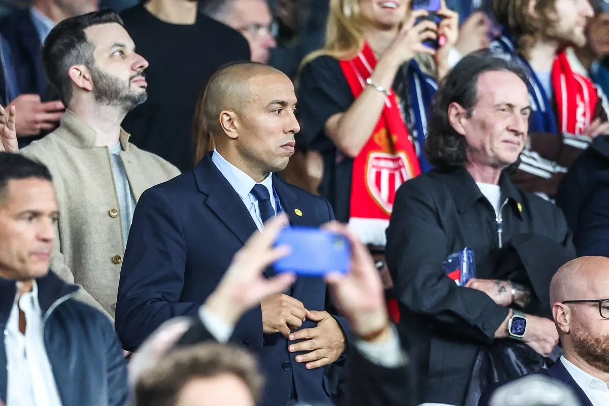 Man in a dark suit stands among cheering spectators at a stadium, hands near his stomach, looking to the left while others watch or film around him.