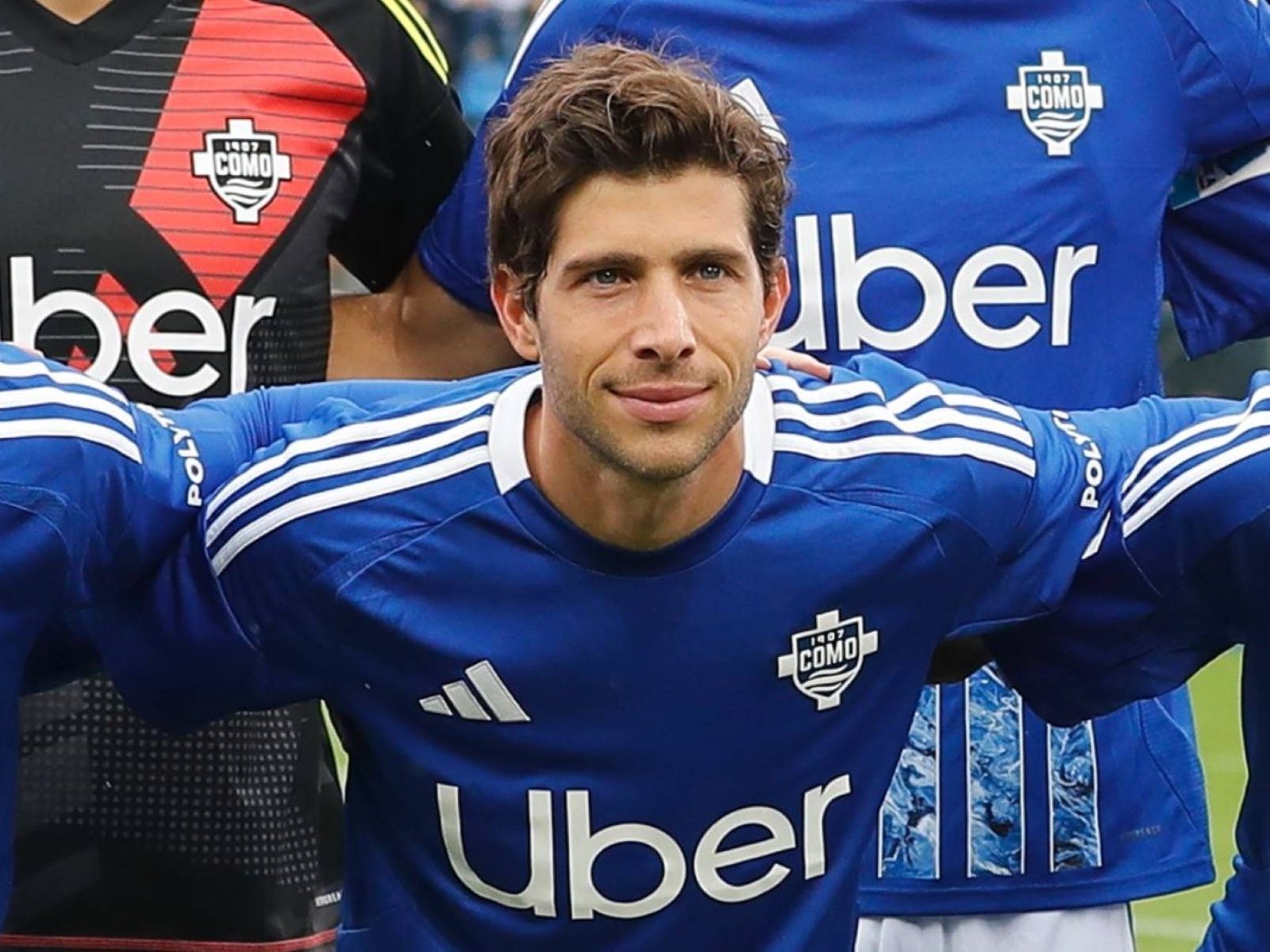 Male soccer player in a blue jersey with Uber logo, posing with teammates in a group photo tourney setting.