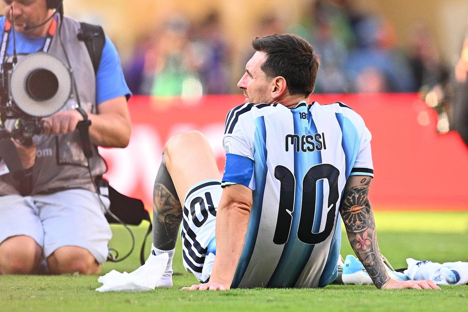 Lionel Messi in an Argentina jersey sits on the grass after a match, while a photographer films nearby in a vest and camera.