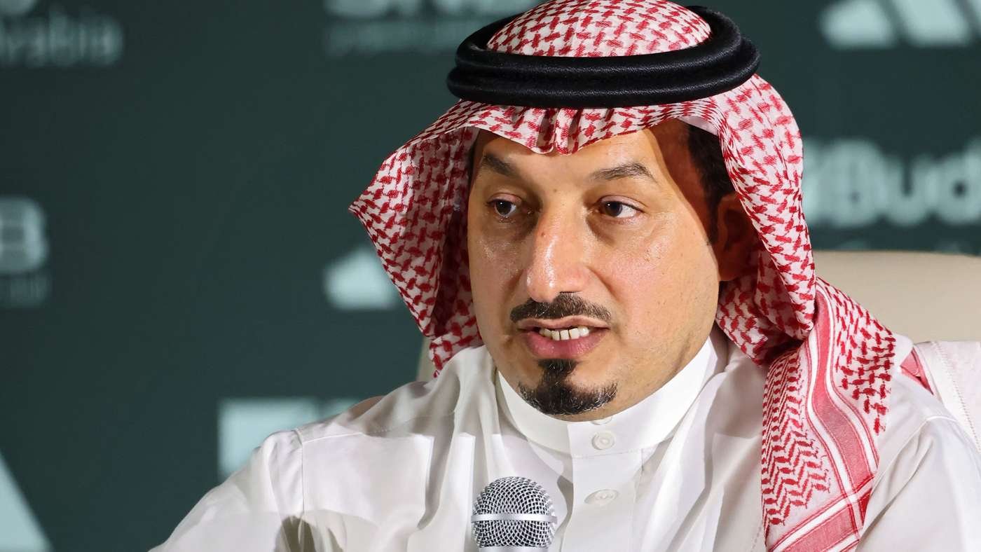 Man in a white thobe and red keffiyeh speaks into a microphone at a conference or panel event at a formal venue.