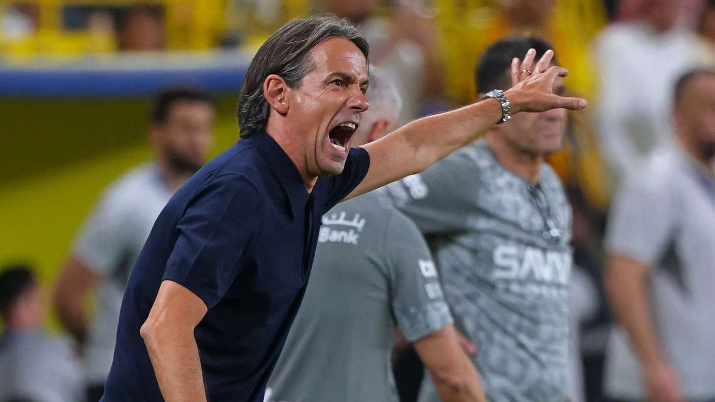 Coach on the sideline shouting with mouth open and arm extended during a game, wearing a dark blue polo shirt.