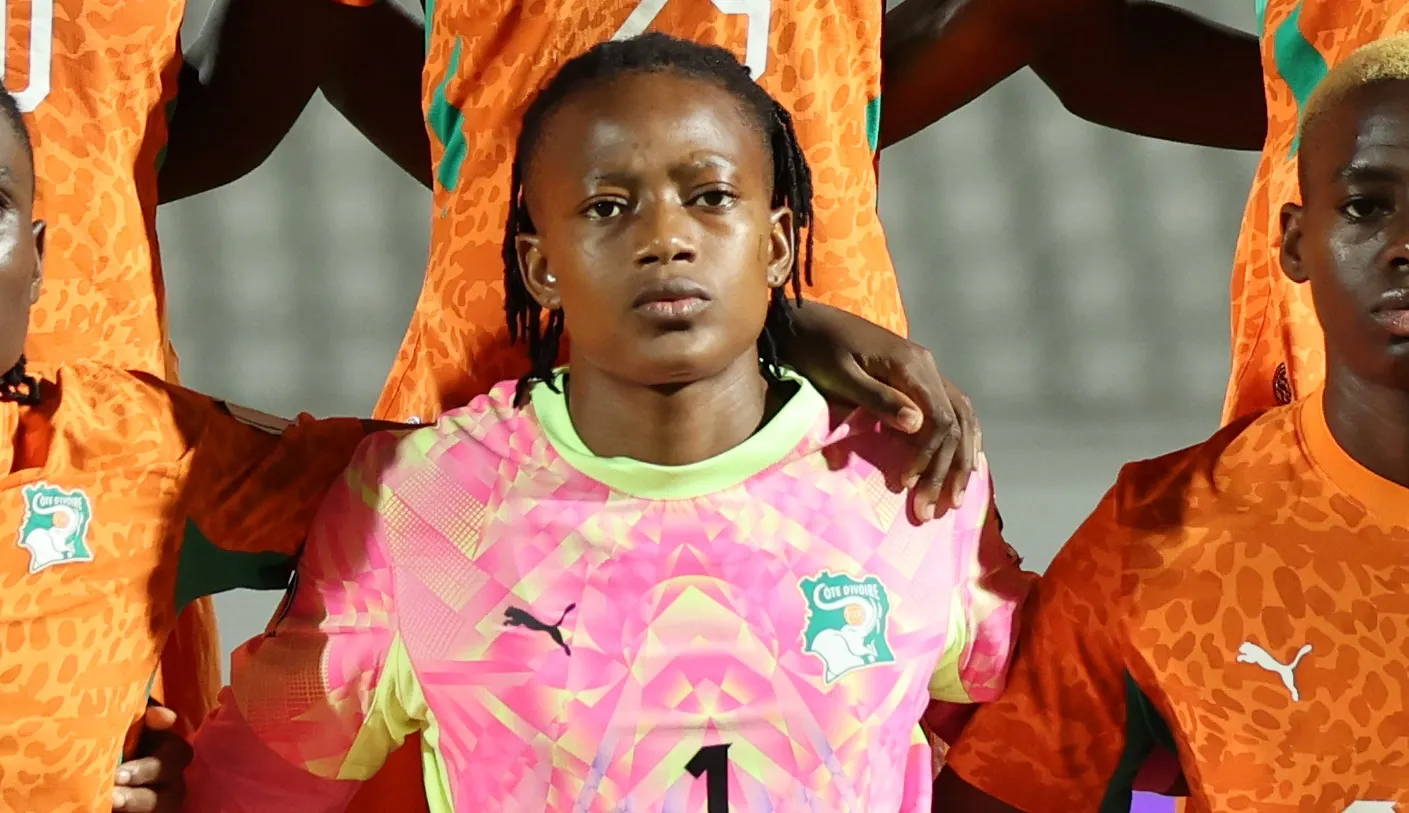 Group of young soccer players with arms around each other; center wears pink goalkeeper jersey, others in orange kits.