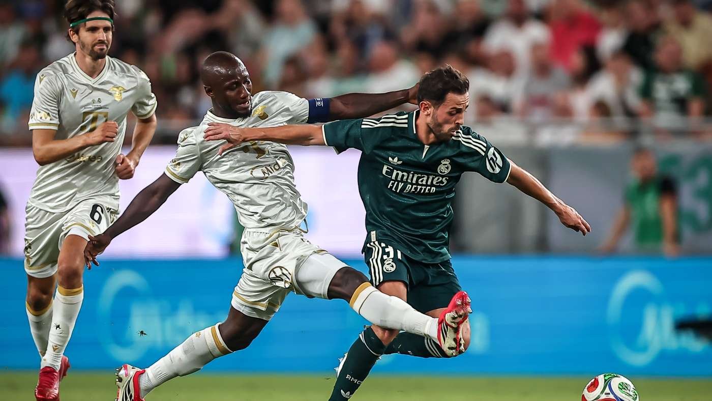 Soccer players from white and dark-green teams clash as a green-clad player kicks the ball while a defender reaches in.