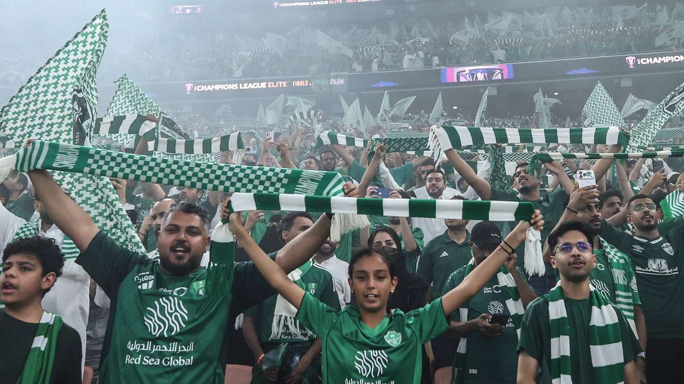 Crowd of green-clad fans in a stadium, holding up green-and-white scarves and cheering.