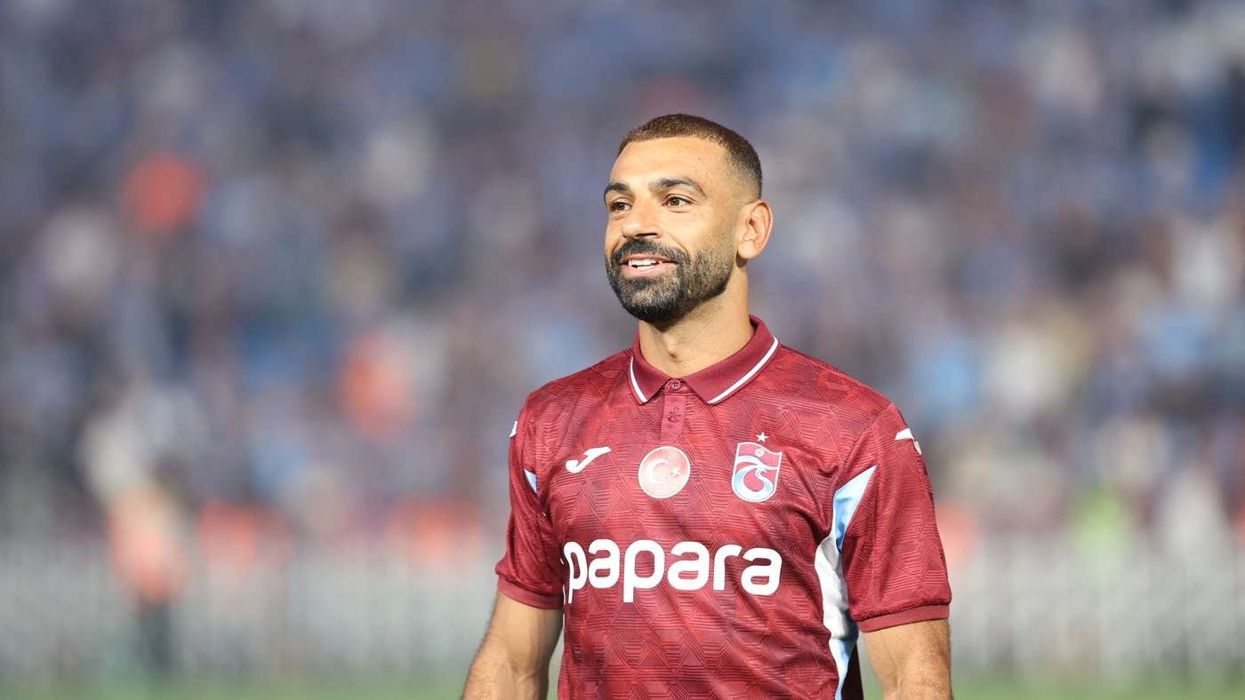 Football player in a burgundy jersey on the pitch, smiling with a blurred stadium crowd behind him.
