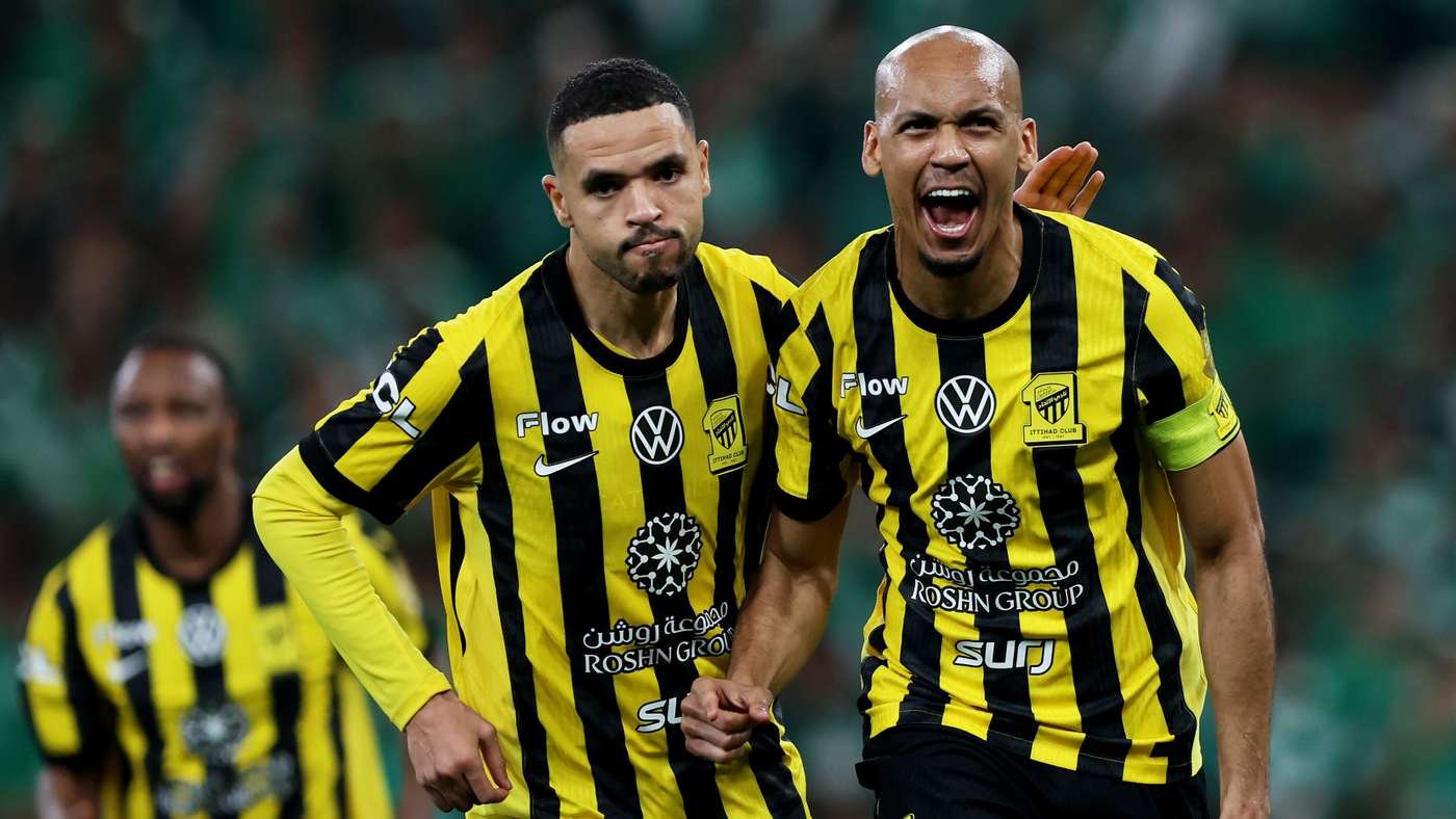 Two soccer players in yellow and black striped jerseys celebrate on the field, fists clenched and expressions of joy, with a blurred green stadium background.