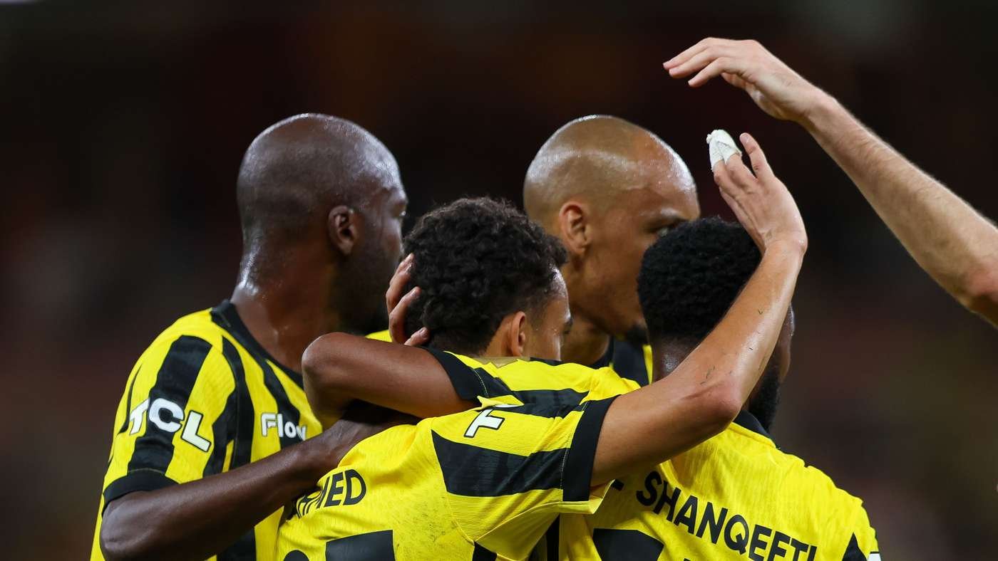 A group of four soccer players in yellow and black jerseys embrace in celebration on the pitch.