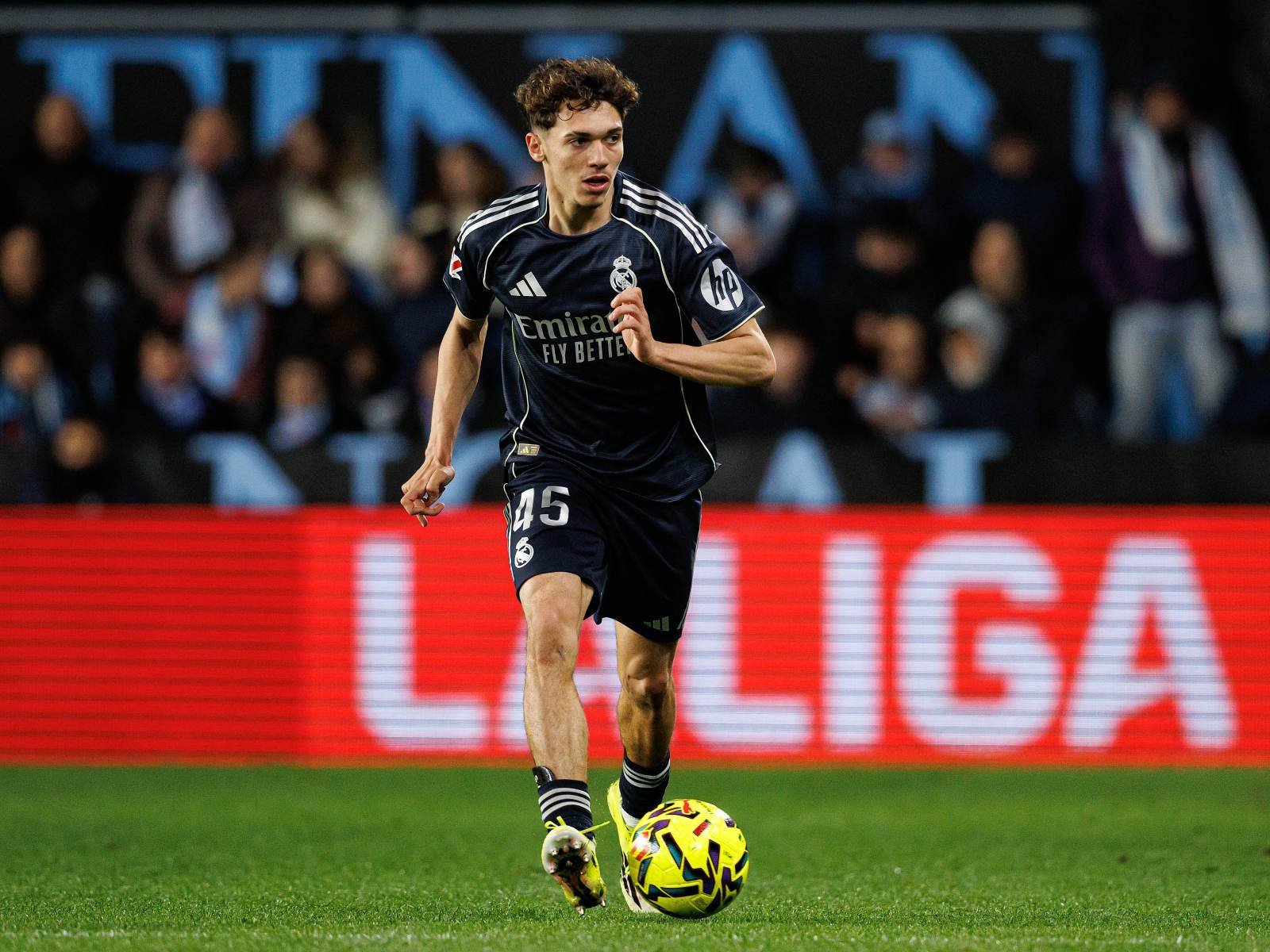 Real Madrid footballer wearing navy kit (#45) dribbles a bright yellow ball on the pitch with spectators in the background.