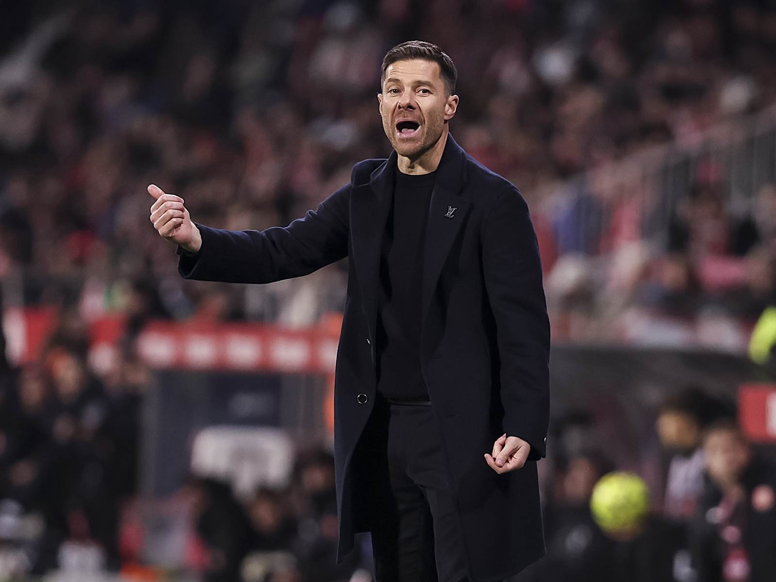 Football coach in a dark coat shouting and gesturing with a clenched fist on the sideline, blurred crowd in the background.