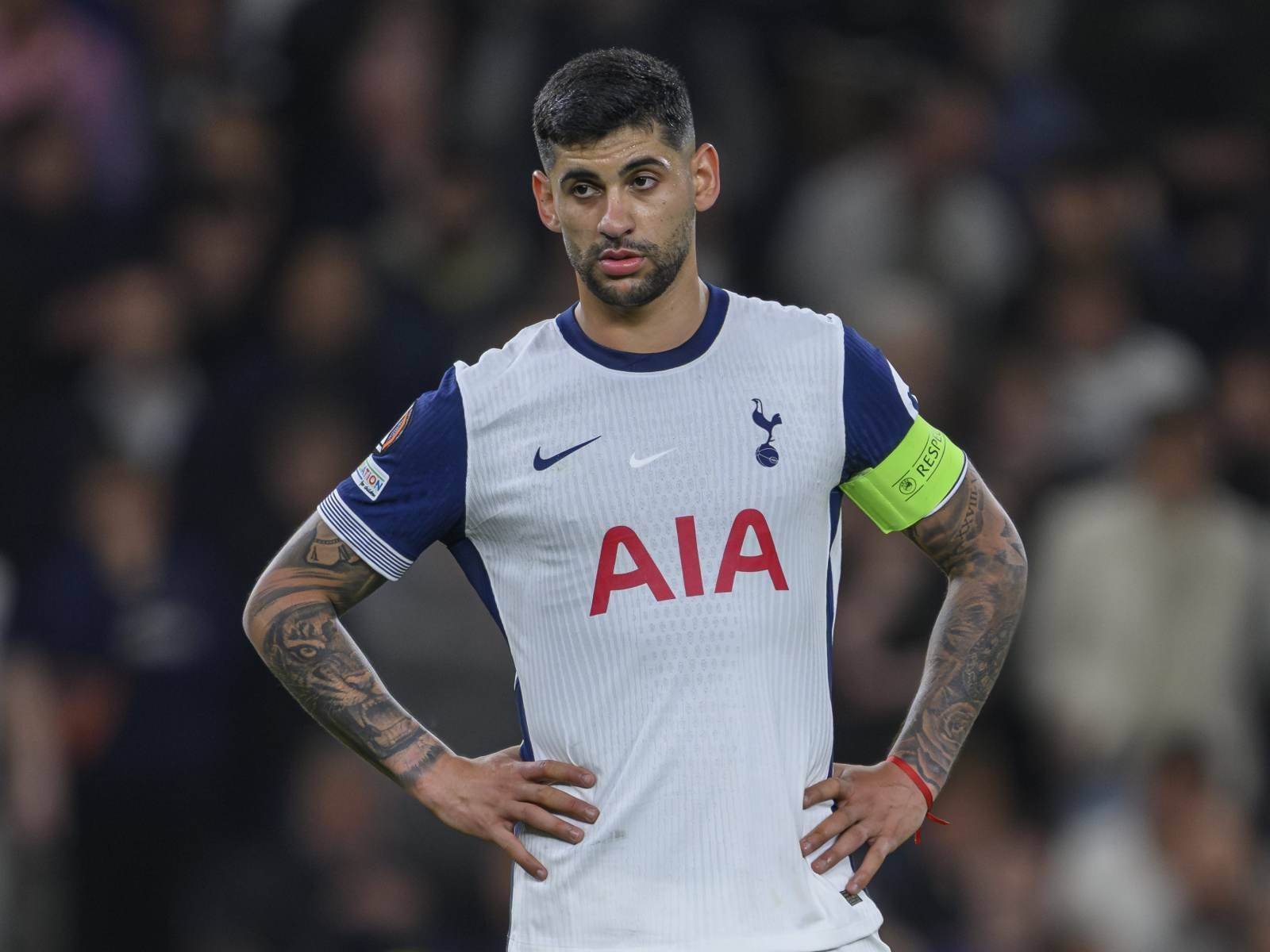Soccer player in a white Tottenham jersey with captain's armband, hands on hips on the field.