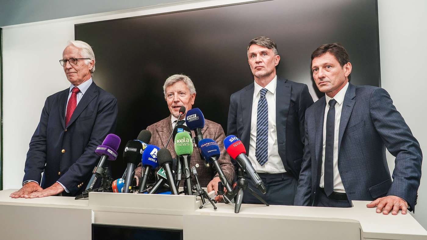 Four men in suits stand behind a white podium at a press conference with many microphones in front of them.