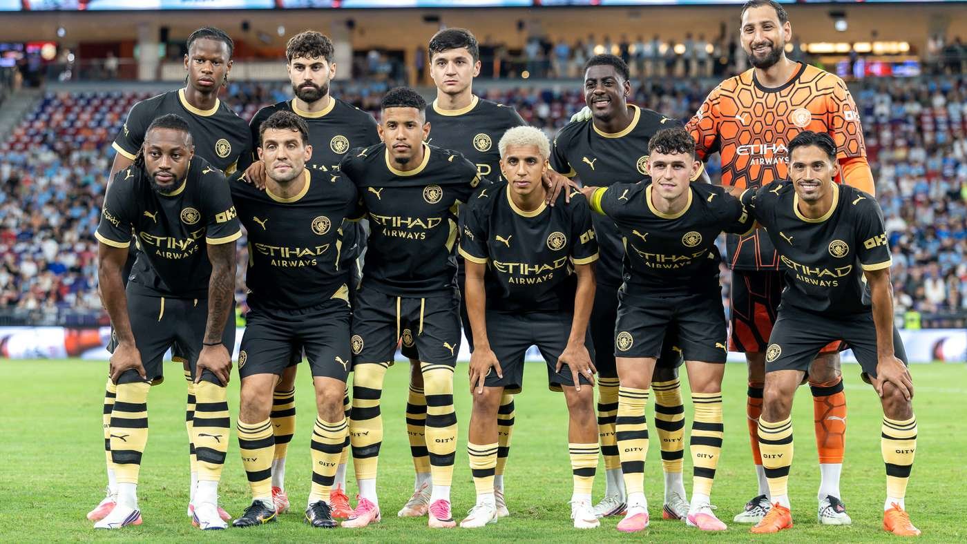 Manchester City players pose for a team photo on the pitch before a match, in black and gold kit with a goalkeeper in orange nearby, stadium in background. Manchester City players pose for a team photo on the pitch before a match, in black and gold kit with a goalkeeper in orange nearby, stadium in background.