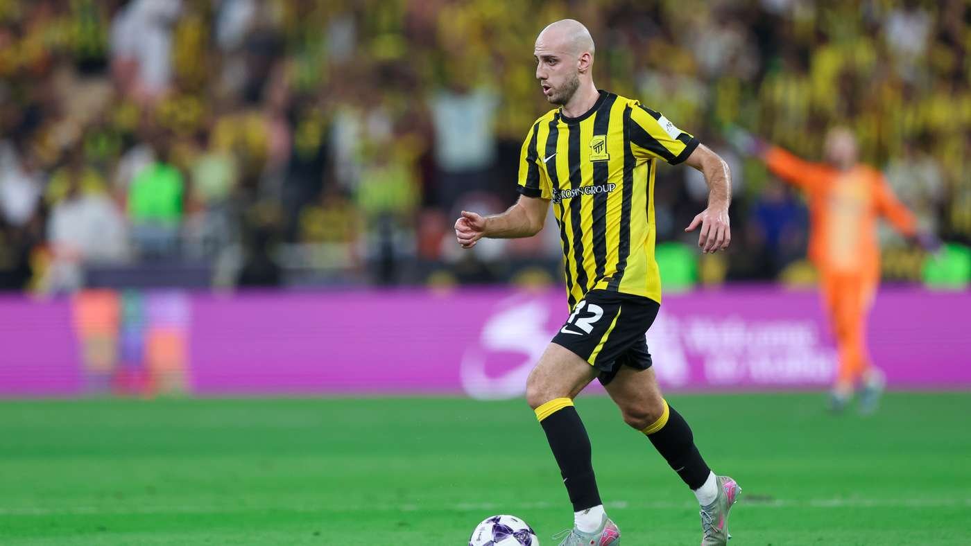 Soccer player in yellow-and-black striped jersey dribbling a ball on a green pitch with a blurred stadium crowd in the background, number 12 on shorts.