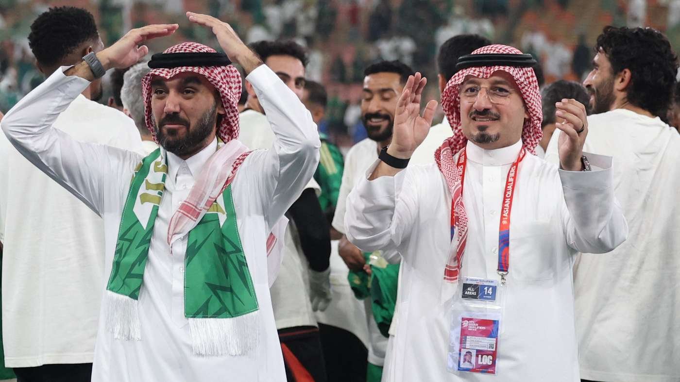 Two men in white thobes and red checkered headscarves celebrate at a sports event, arms raised.