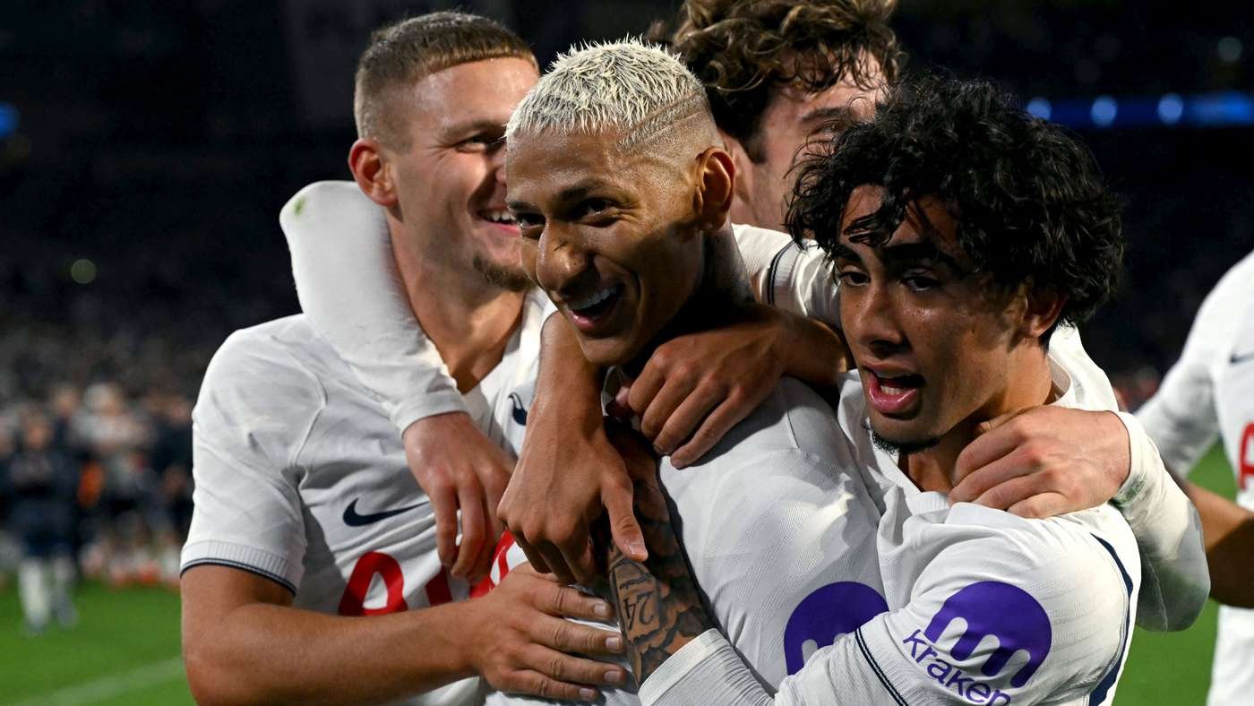 Group of soccer players in white jerseys celebrating with arms around each other on a green field at night.