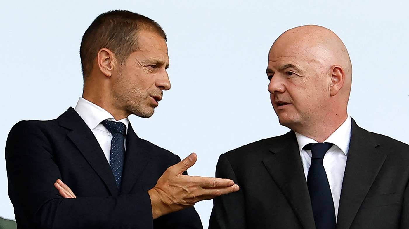 Two men in dark suits converse, one gesturing with his hand toward the other against a pale blue sky.