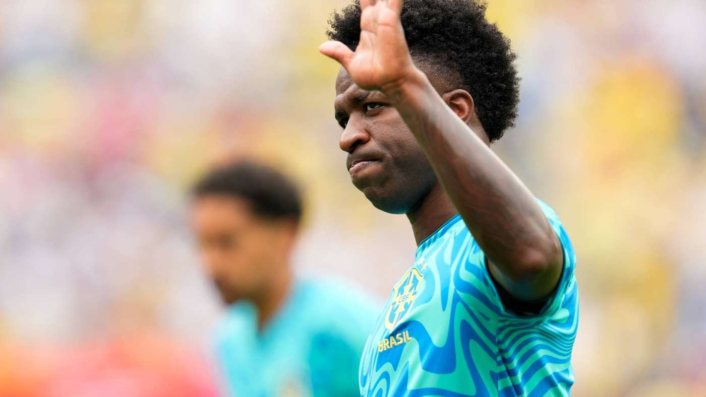 Soccer player in Brazil jersey raises hand in gesture on the field with a blurred opponent in the background.