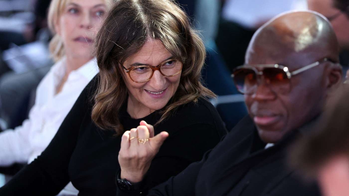 Woman with glasses smiles and looks at a man wearing sunglasses in a crowded setting, her hand raised near her face. Woman with glasses smiles and looks at a man wearing sunglasses in a crowded setting, her hand raised near her face.