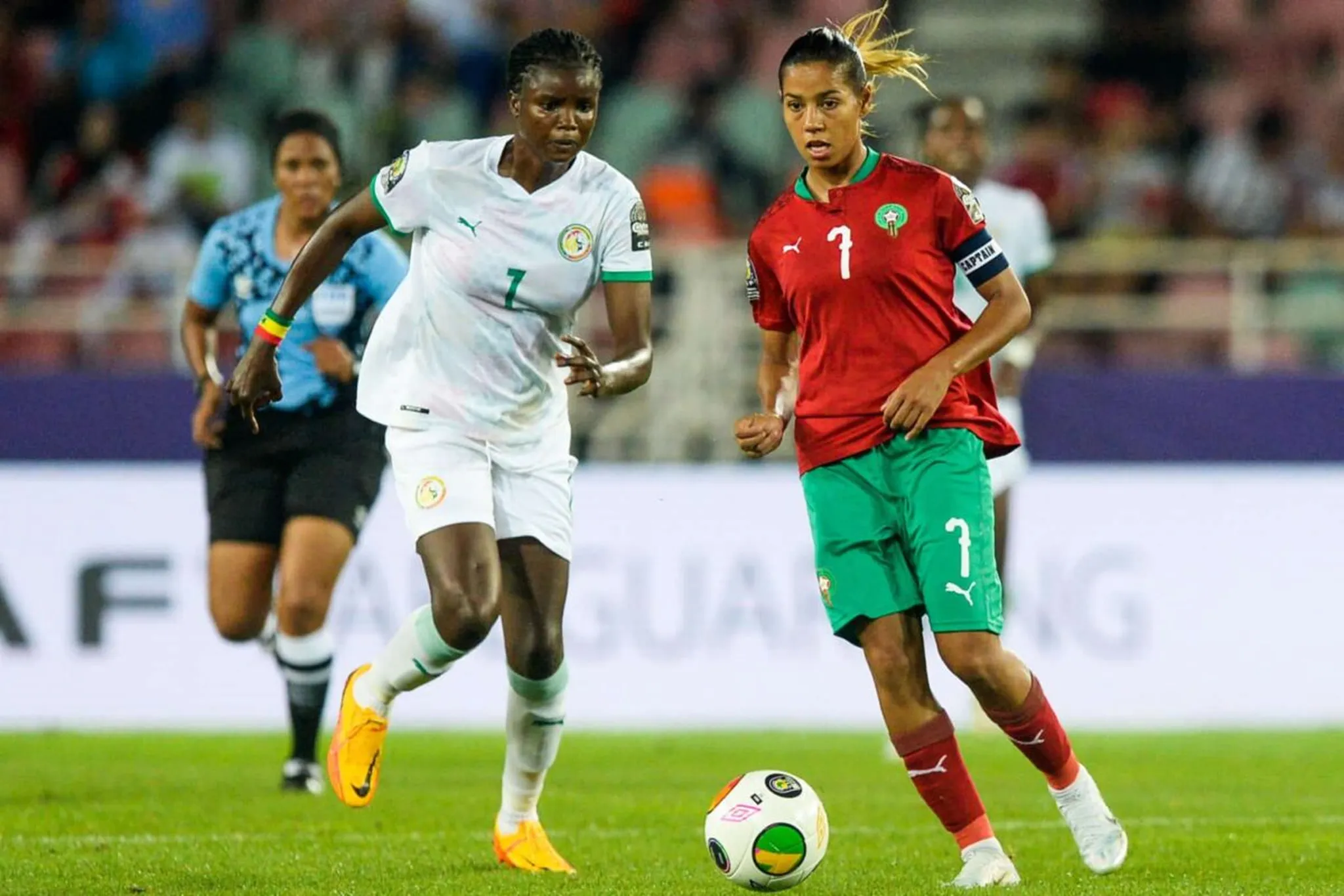 Two female soccer players compete for the ball on a green field, one in white uniform (number 7) and one in red (number 1) with a captain's armband on her sleeve.