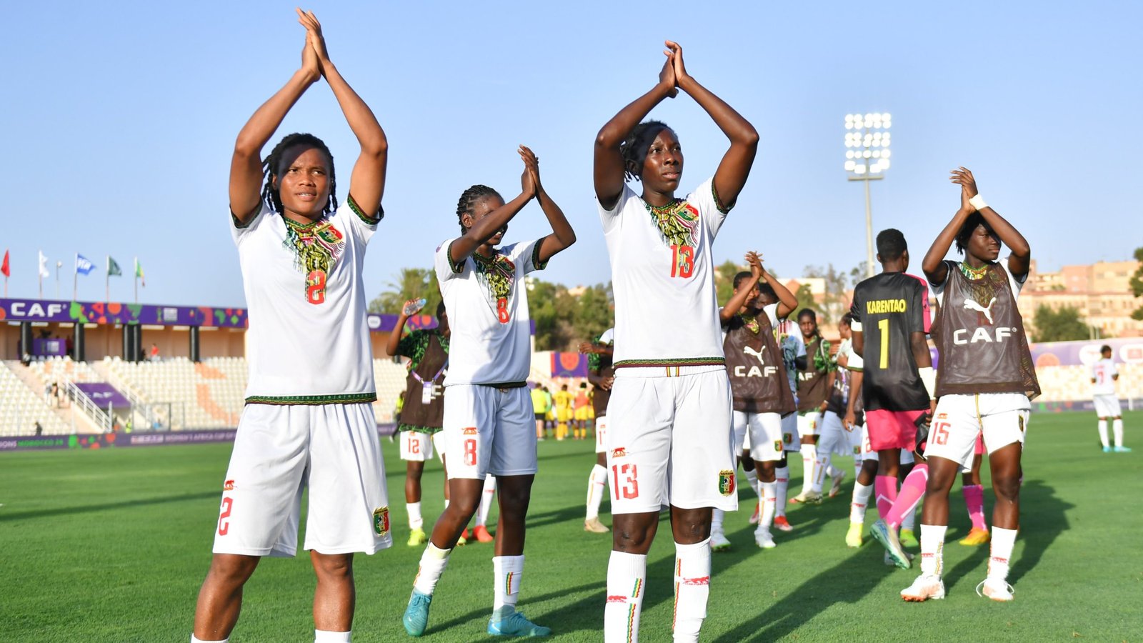 Soccer players in white jerseys raise their hands together in a prayer-like pose on a sunlit field after a game in a stadium