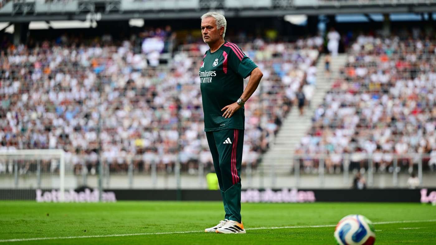 Football manager standing on a green pitch with hands on hips, a full stadium behind him.