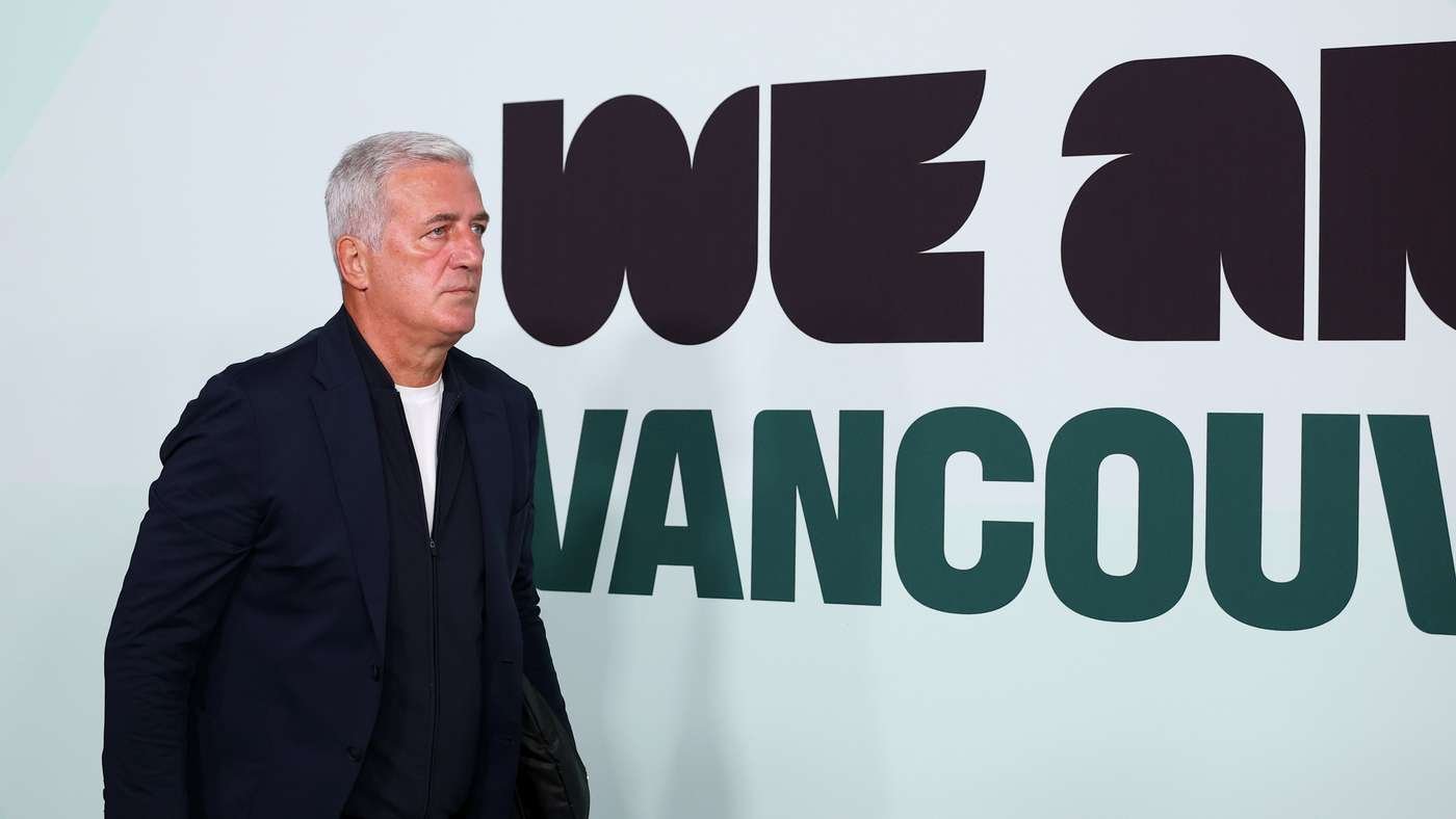 Middle-aged man in a dark blazer stands in front of a wall with large bold lettering in black and green.