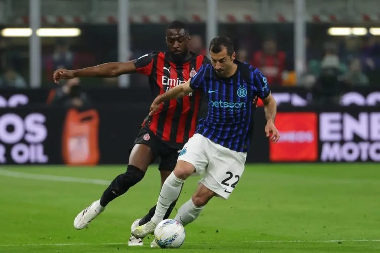 Two soccer players in red-and-black and blue-and-black kits contest a ball on the field during a match.