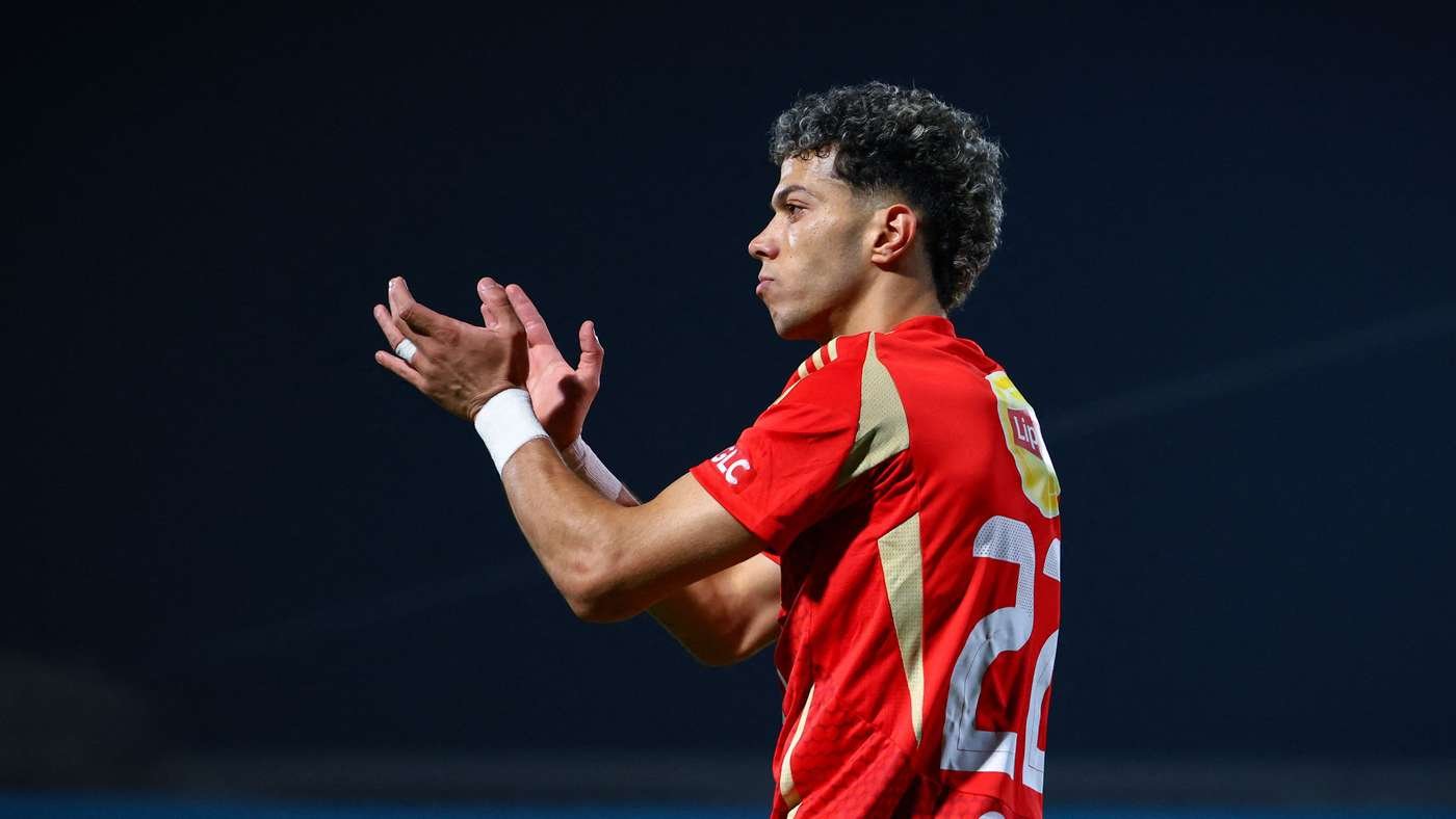 Soccer player in a red jersey applauding on the field with a dark background behind him.
