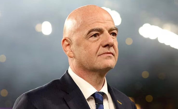 Bald man in a dark suit and blue tie, looking to the right, with soft bokeh lights in the background.