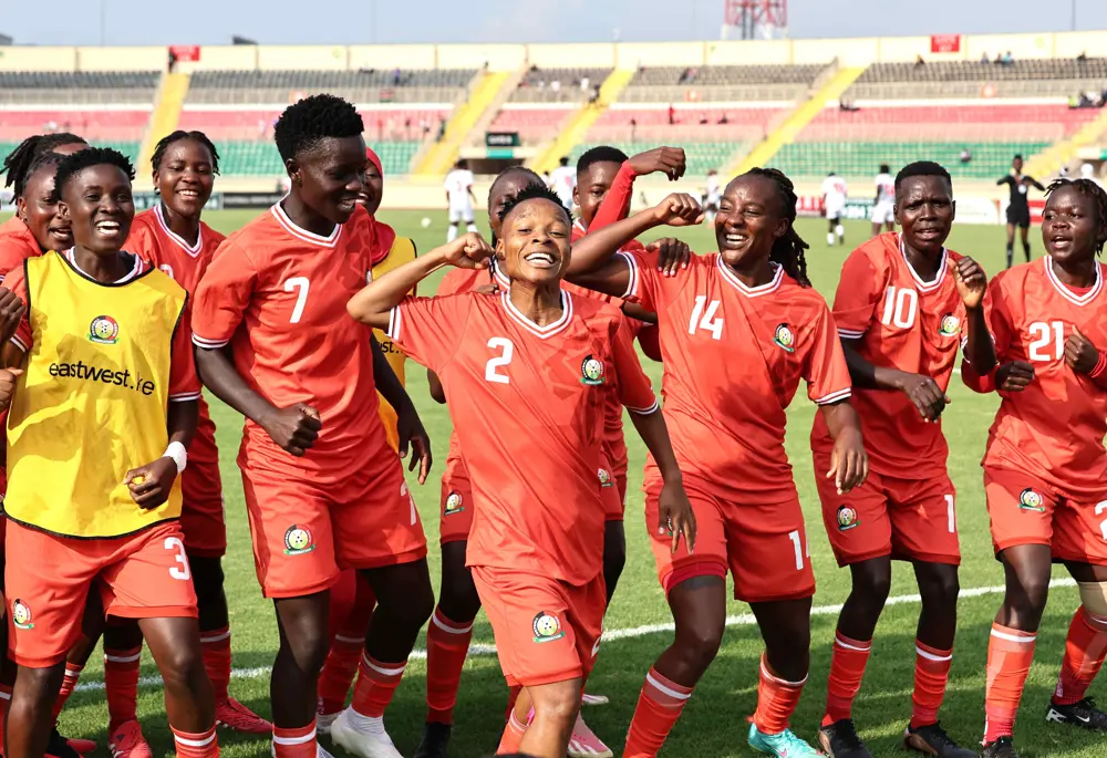 Group of female soccer players in red uniforms celebrate together on a sunny stadium field; spectators sit in the stands behind them.