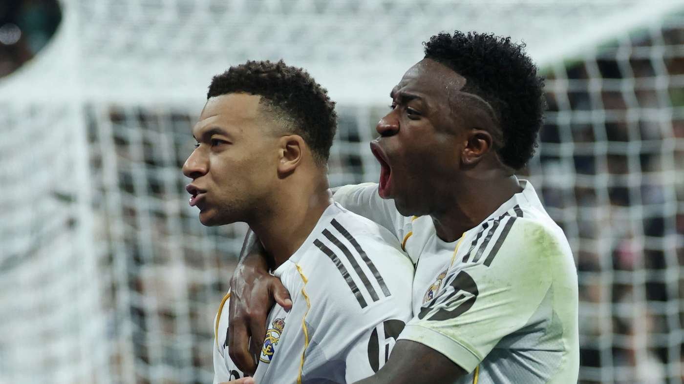 Two soccer players in white jerseys celebrate a goal, one hugging the other from behind in front of a net.