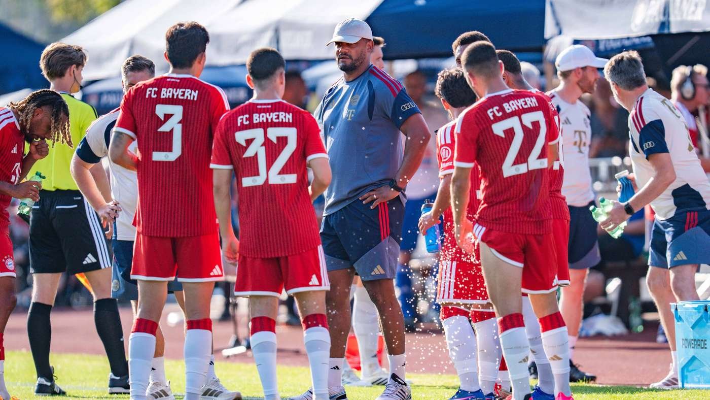 Soccer players in red FC Bayern jerseys stand in a circle around a coach in a blue shirt during a break on the field, water bottles in hand.