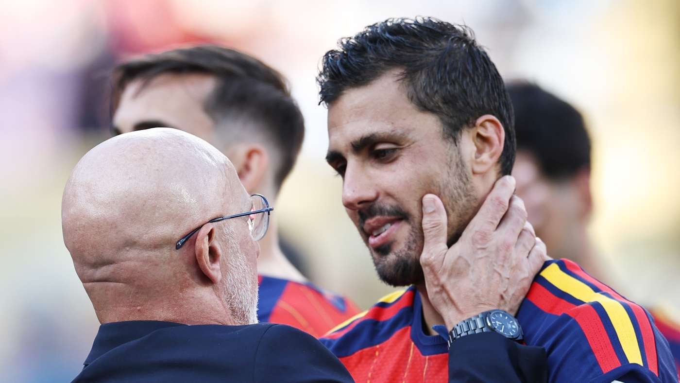 Two men in red and blue sports jerseys share a close, supportive moment; the older man with glasses touches the younger man's face as they converse on a field. Two men in red and blue sports jerseys share a close, supportive moment; the older man with glasses touches the younger man's face as they converse on a field.