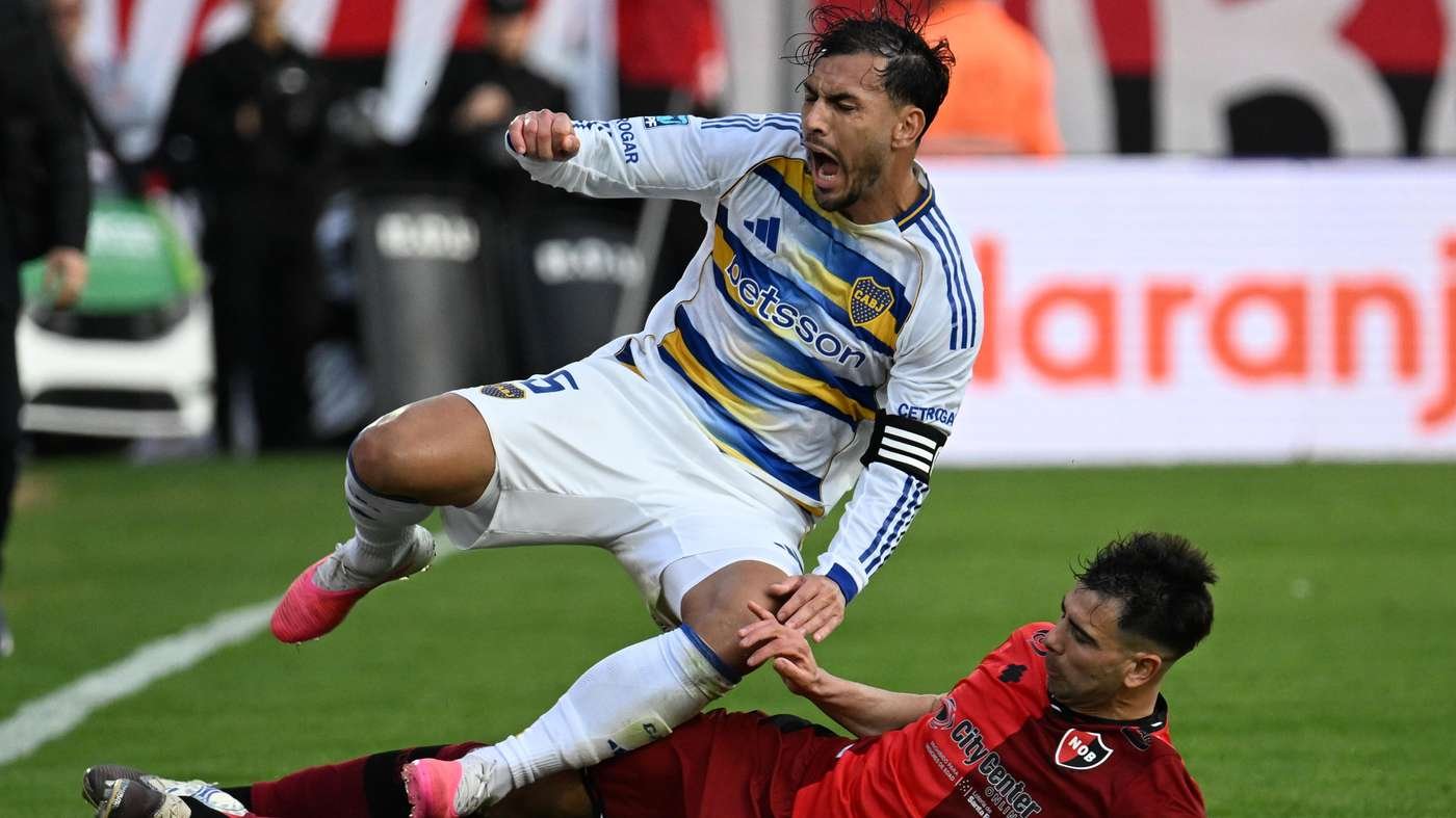 Two soccer players in a sliding challenge during a match, one in white-blue kit, the other in red, on a green field with advertising boards in the background Two soccer players in a sliding challenge during a match, one in white-blue kit, the other in red, on a green field with advertising boards in the background