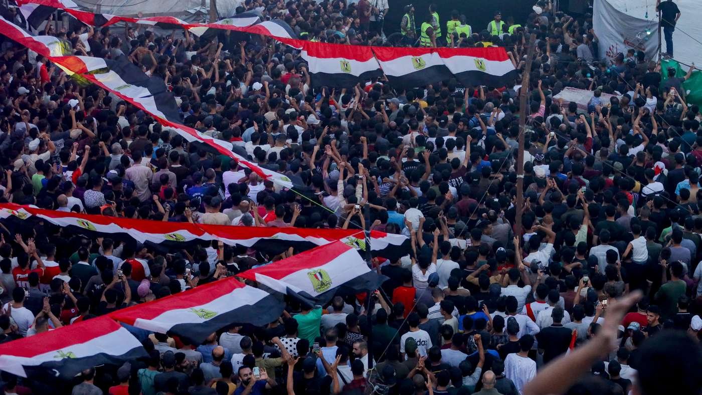 Large crowd at a protest, many people waving Egyptian flags and raising their hands.