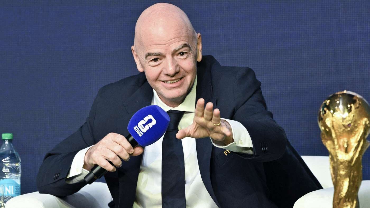 Bald man in a dark suit speaks into a blue microphone during a press event, with a golden trophy visible on the right and a water bottle on the left.