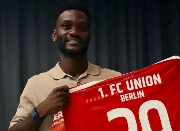 Smiling man in a beige polo shirt holds up a red 1. FC Union Berlin scarf showing the number 20 against a dark backdrop.