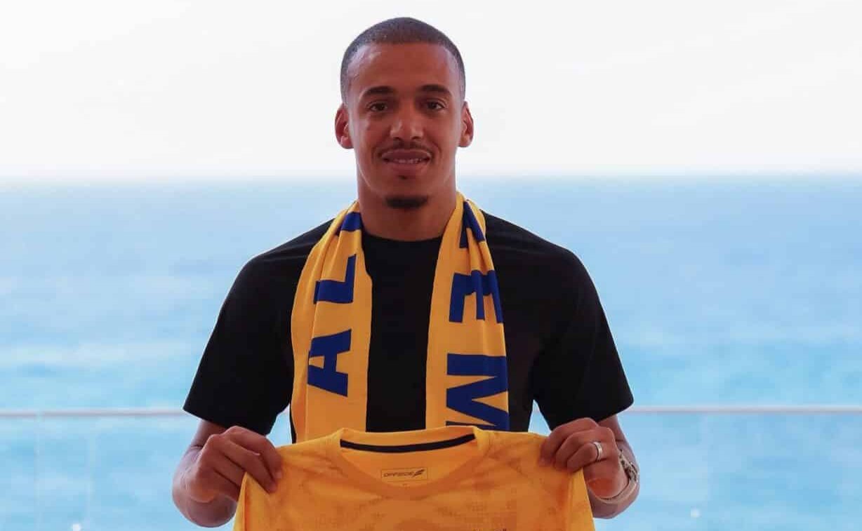 Man with short hair wearing a black shirt, yellow scarf, and holding a yellow jersey by the sea.