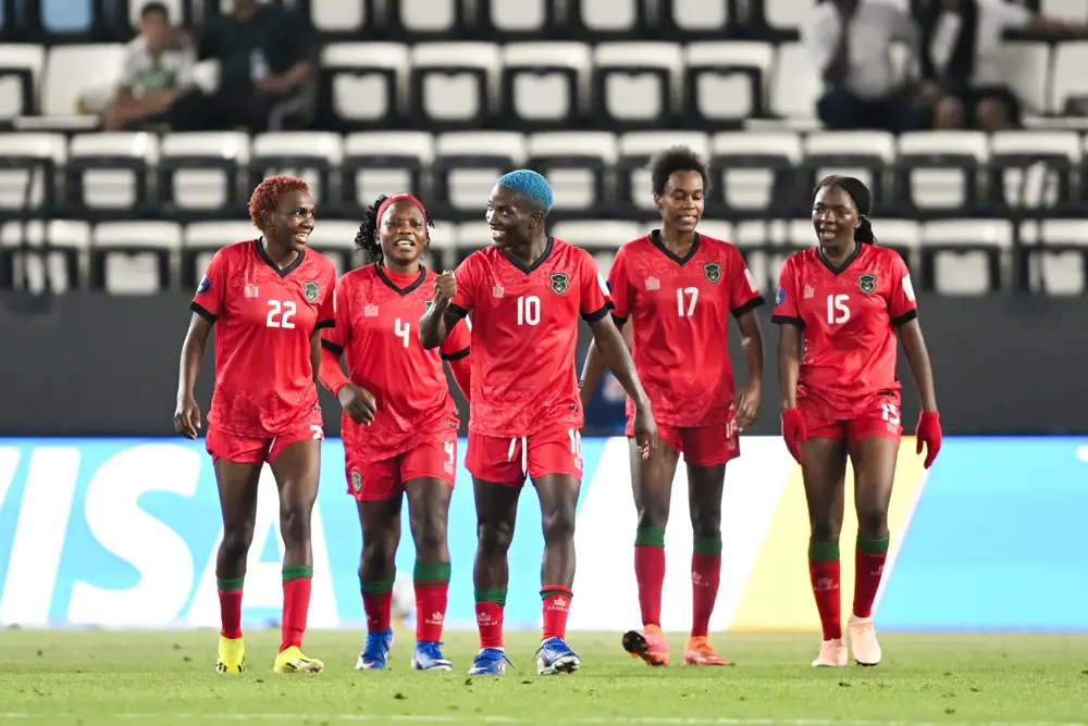 Five female soccer players in red uniforms walk together on a football pitch, smiling and chatting.