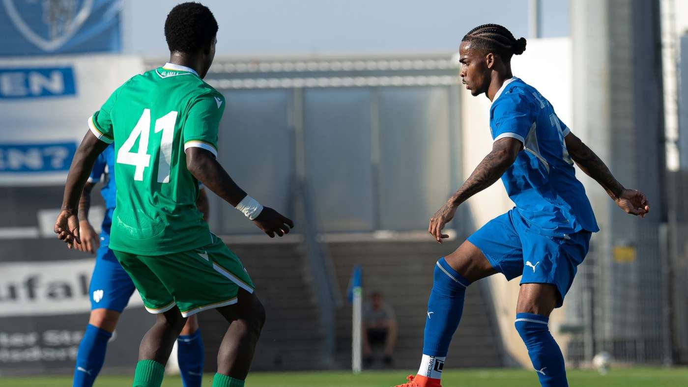 Two soccer players in green and blue kits compete for the ball on a sunny field.
