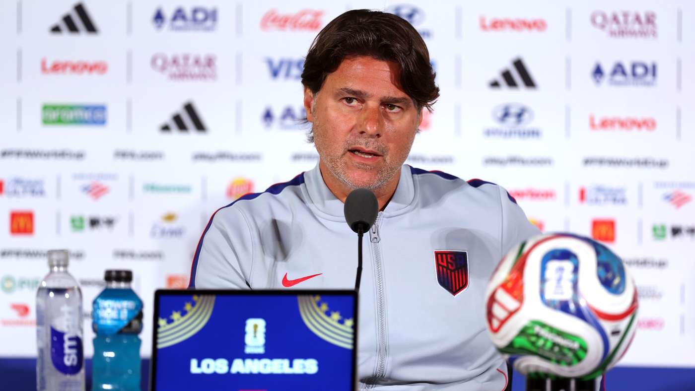 Man in a gray USA soccer jacket speaks into a microphone at a press conference, with a colorful soccer ball beside him and sponsor backdrop behind.