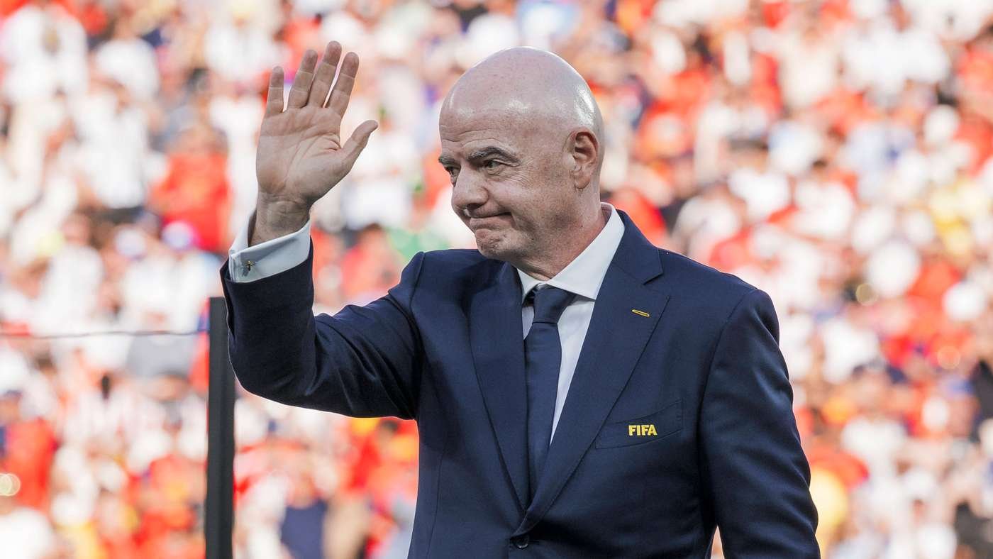 Bald man in a navy suit waving to a blurred crowd at a stadium, FIFA logo on his lapel.